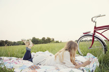 young woman reading on a blanket in the grass, next to her bicycle 