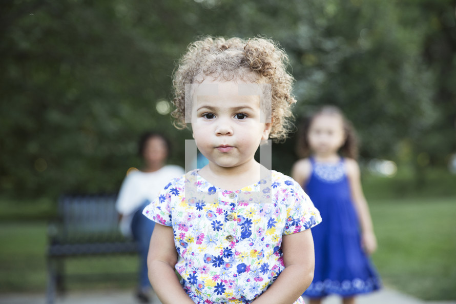 toddler girl standing in front of her family sitting on a park bench 