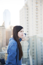 a woman leaning on a glass wall on a rooftop balcony thinking 