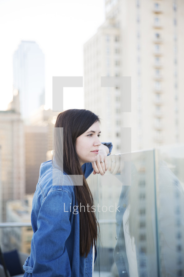 a woman leaning on a glass wall on a rooftop balcony thinking 