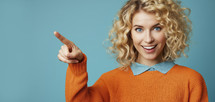 Joyful young woman with blonde curly hair, pointing finger, wearing an orange sweater and blue collar, with a vibrant teal background.