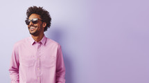 Smiling young man with curly hair wearing aviator sunglasses and a light pink shirt on a lavender background.