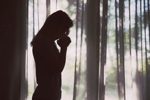 silhouette of a woman with head bowed in prayer standing alone in a forest 