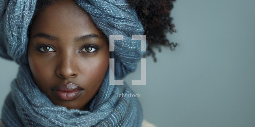 Portrait of a young woman with a serene expression, wrapped in a soft denim-textured scarf, set against a pale blue backdrop.