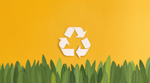 Recycling symbol with three arrows on a yellow background above green grass, representing environmental conservation and sustainability.