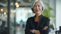 Sophisticated mature businesswoman with silver hair, arms crossed, and a self-assured smile in a modern corporate environment.