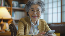 Elderly Asian woman with phone, cozy home interior, warm smile.