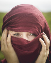a young woman with her head wrapped in a burgundy scarf, only her eyes showing, standing outdoors