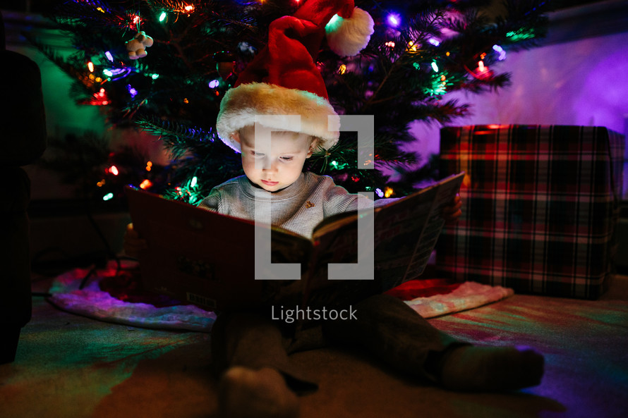 a kid reading a book under a Christmas tree 