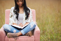 a young woman sitting in a chair in a field reading a Bible and praying 