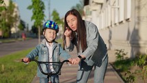Happy family walk in outdoor a parent mother teaches her son child to ride a bike on city street. Woman happy supports child and rejoices the success of boy. Sports walk in nature. Healthy lifestyle.