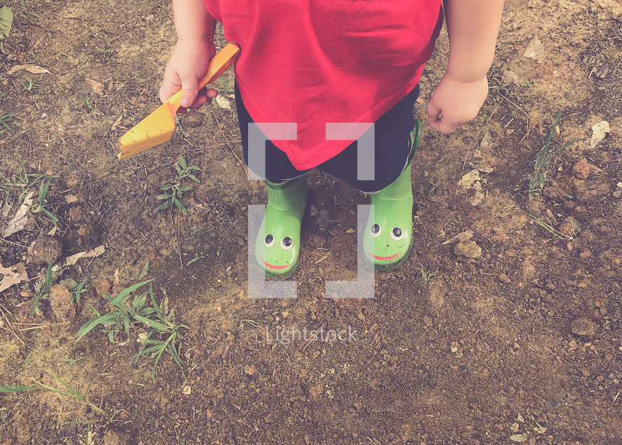 a toddler boy in rain boots playing in dirt 