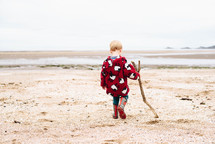 a boy child in rain boots dragging a stick on a beach 