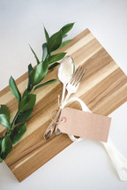 A fork and spoon and a sprig of green leaves on a wooden cutting board.
