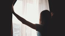 Beautiful woman opening curtains and looking through the window. Sunrise or sunset behind the window. Young woman opening curtains in a bedroom. Positivity and energy concept.
