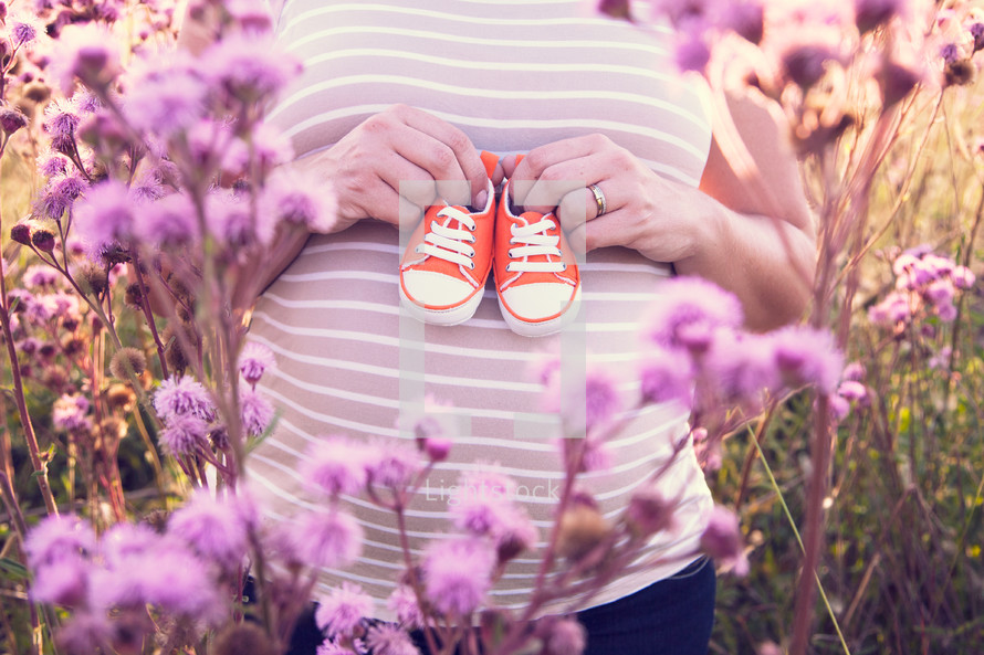 Pregnant woman holding a pair of baby shoes in a field of flowers.