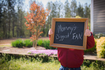 toddler boy holding a chalkboard sign that reads mommy's biggest fan 