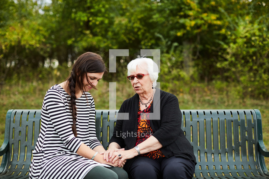 elderly woman praying with a young woman on a park bench 