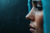 Close-up of a serene face under a hood in the rain, highlighting water droplets on the skin, evoking introspection and calm.