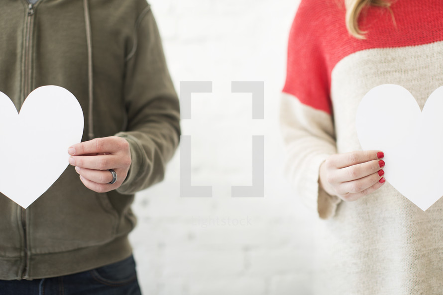 A man and woman stand side by side holding white paper hearts.