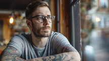 Pensive man in cafe, tattooed arms, trendy glasses.