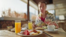 Young woman eats breakfast in the wooden cafe on mountain, alpine view, snow on hills. Breakfast on terrace of cafe in mountain resort. Healthy lifestyle, travel, family moments together concept.