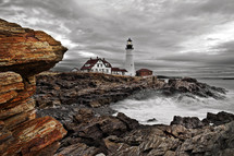 Portland Headlight Lighthouse In Cape Elizabeth Maine. Bright orange rocks in the foreground and a grey overcast sky.