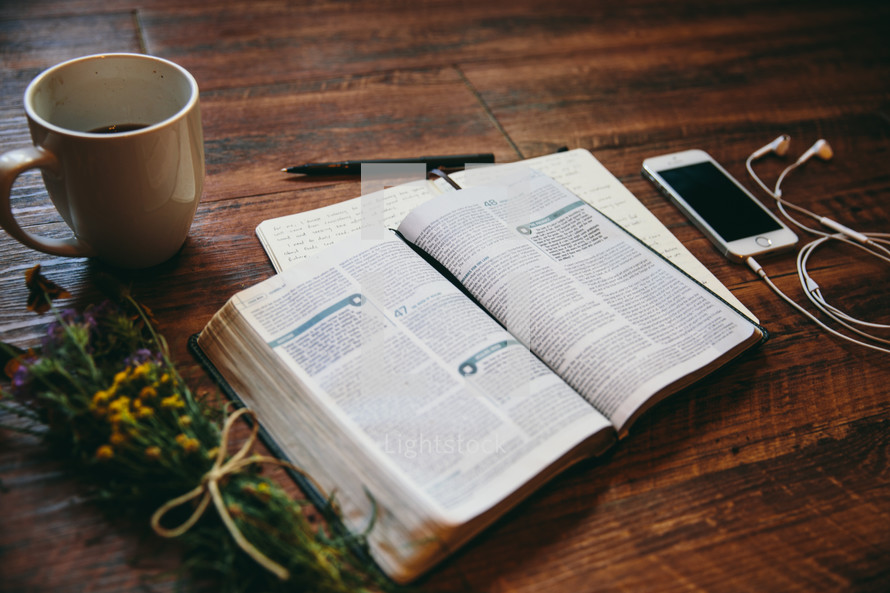 open Bible, coffee mug, iPhone, journal, earbuds, flowers, and a pen on a table