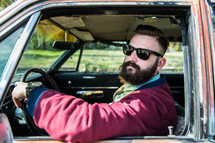 A man with a beard and sunglasses driving a car.