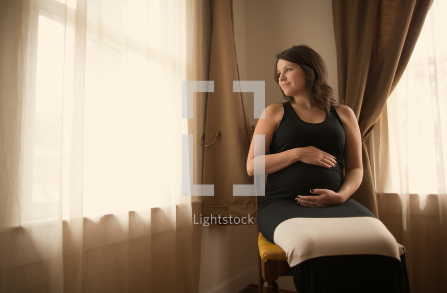 pregnant woman sitting in a chair holding her belly looking out a window