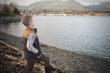 Young boy child in boots standing on a rocky shore pondering his life. 