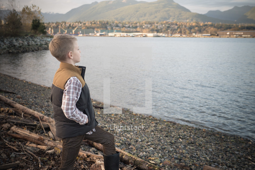 Young boy child in boots standing on a rocky shore pondering his life. 