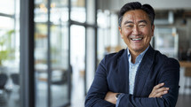Cheerful mature Asian businessman with crossed arms, confidently smiling in a modern office setting.