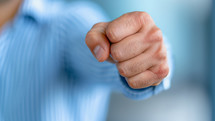 Close-up of a clenched fist in a forward motion, symbolizing determination, strength, and assertiveness, with a person wearing a blue-striped shirt in the background.