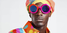 Vibrant portrait of a young man with bold fashion sense, wearing colorful headwear and oversized pink sunglasses against a white backdrop.