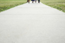 Family’s feet walking down a sidewalk path. 
