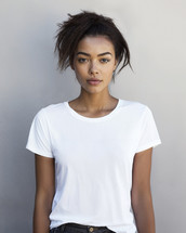 A young, ethnically diverse woman with a natural look confidently poses in a plain white tee, her gaze direct and engaging against a subtly textured urban wall.