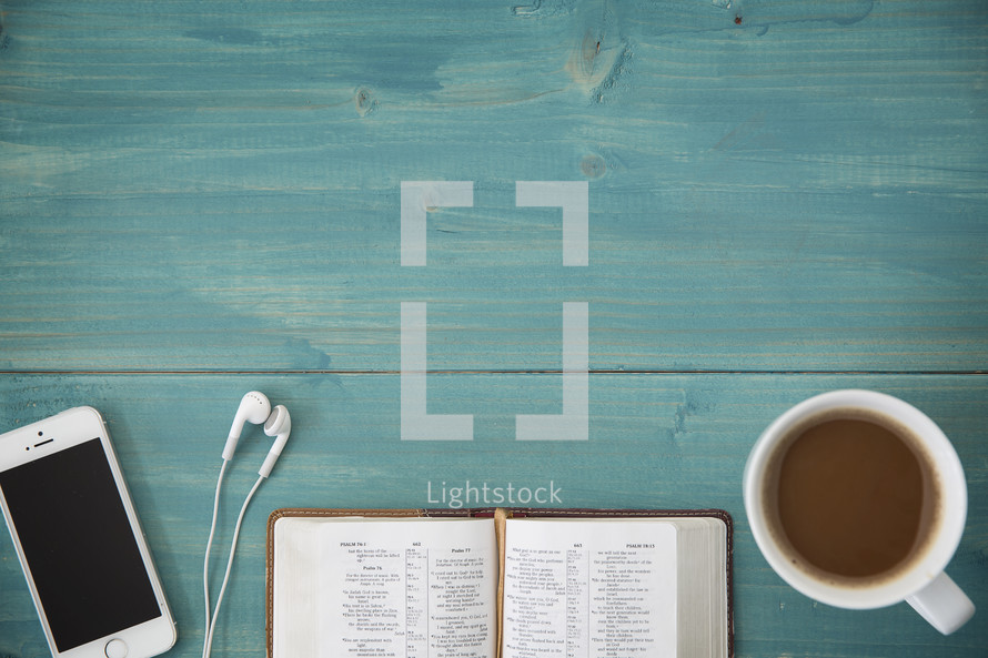 An open Bible, cellphone, ear buds, and cup of coffee on a green wooden table.