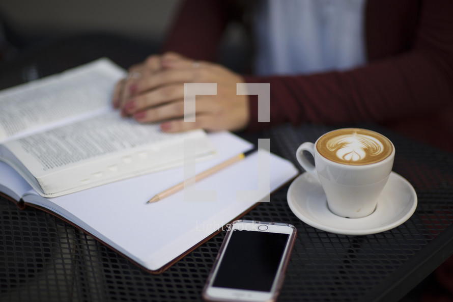 a young woman reading a Bible and journaling at a table with a mug of coffee and cellphone 
