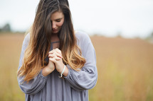 a woman with head bowed and praying hands standing in a field praying 