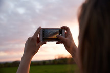 a woman taking a picture of the sky at sunset with her smartphone 