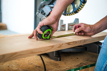 Man holding a measuring tape over a piece of wood under a chop saw.