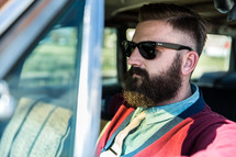 A man with a beard and sunglasses sitting in a car.