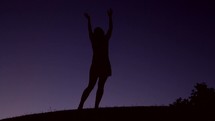 Silhouette of a woman dancing on a hill and raising her hands in praise at night.
