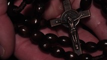 A man's hands holding a wooden Rosary comes out of the dark into the light.