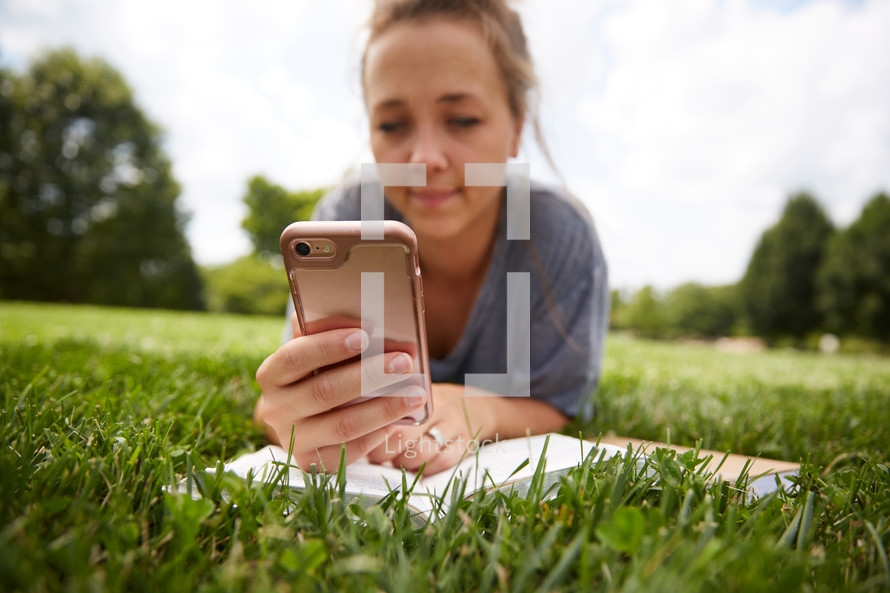 a person sitting in the grass reading a Bible and checking her cellphone 