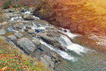 Male hiker with backpack walking on rocks and waterfall in forest, autumn environment