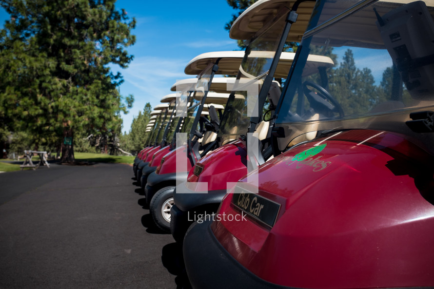 Row of golf carts on asphalt near trees.