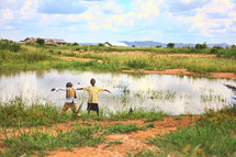 Boys with arms spread standing on the edge of a pond by a village, looking toward the sky.