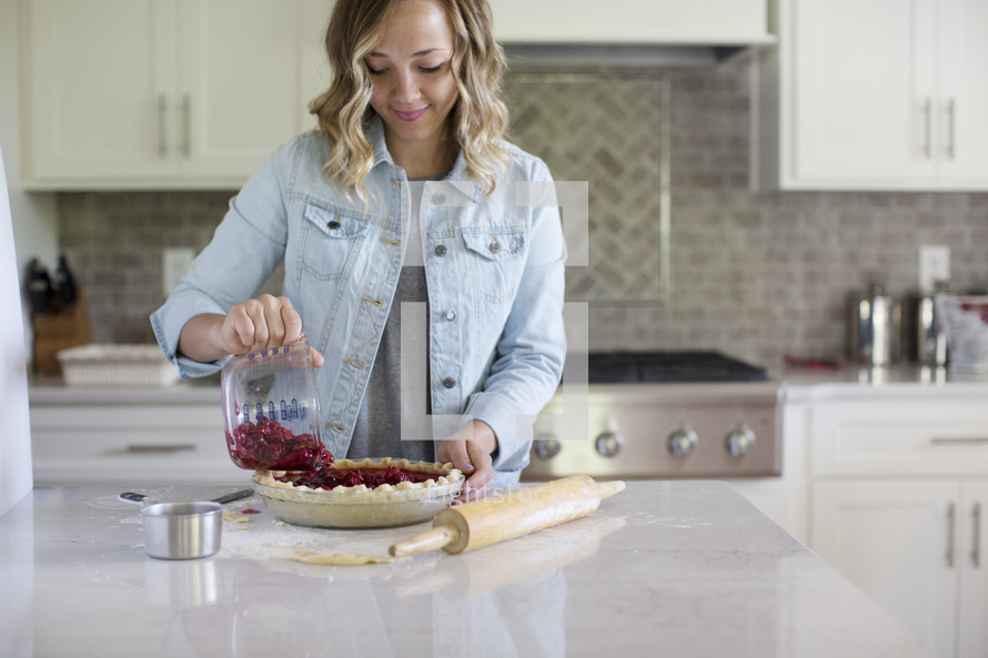 A woman in a kitchen pouring cherries into a prepared pie crust.